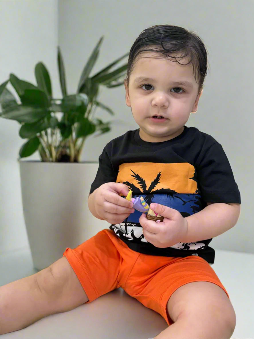 Child wearing a black and orange shirt with a colorful design, sitting on a bed with a white and green patterned pillow.