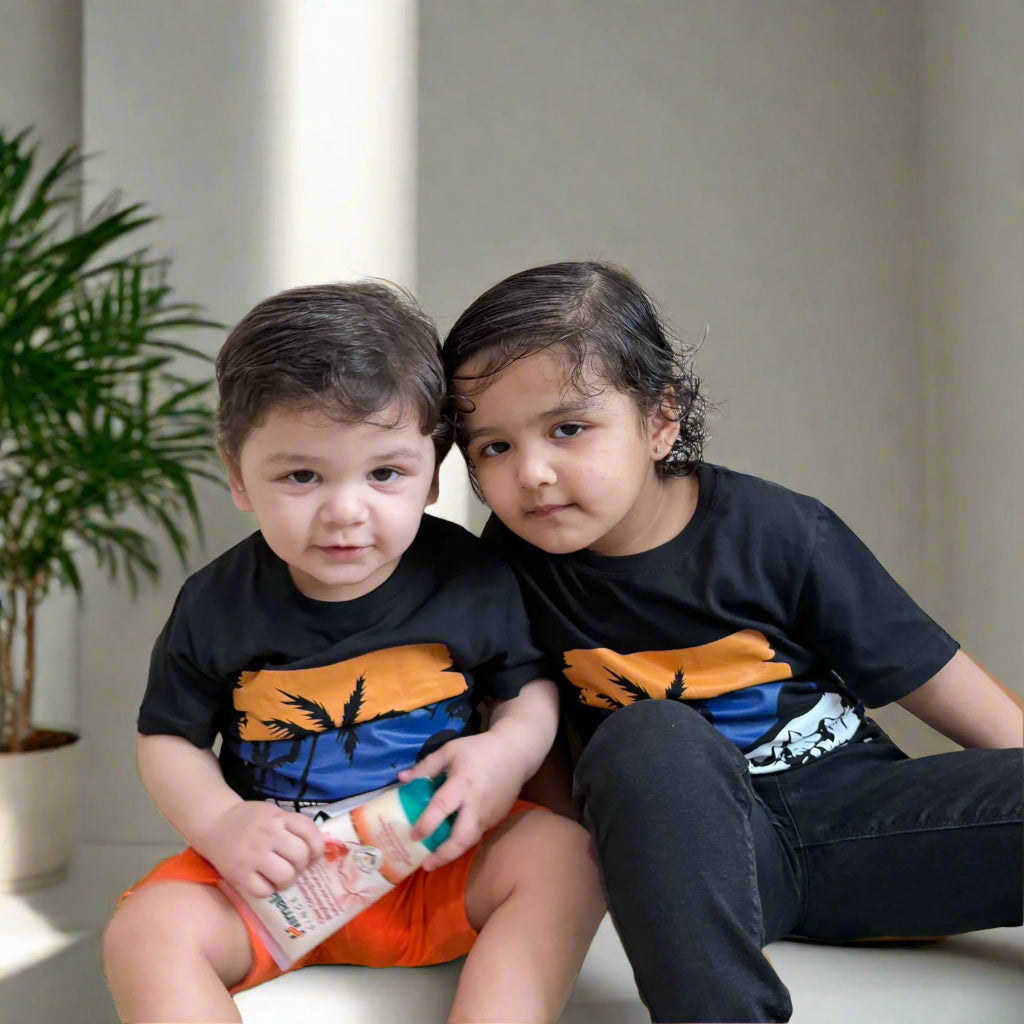 Two children wearing matching black t-shirts with orange and blue designs, sitting on a white surface.
