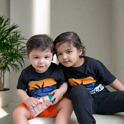 Two children wearing matching black t-shirts with orange and blue designs, sitting on a white surface.