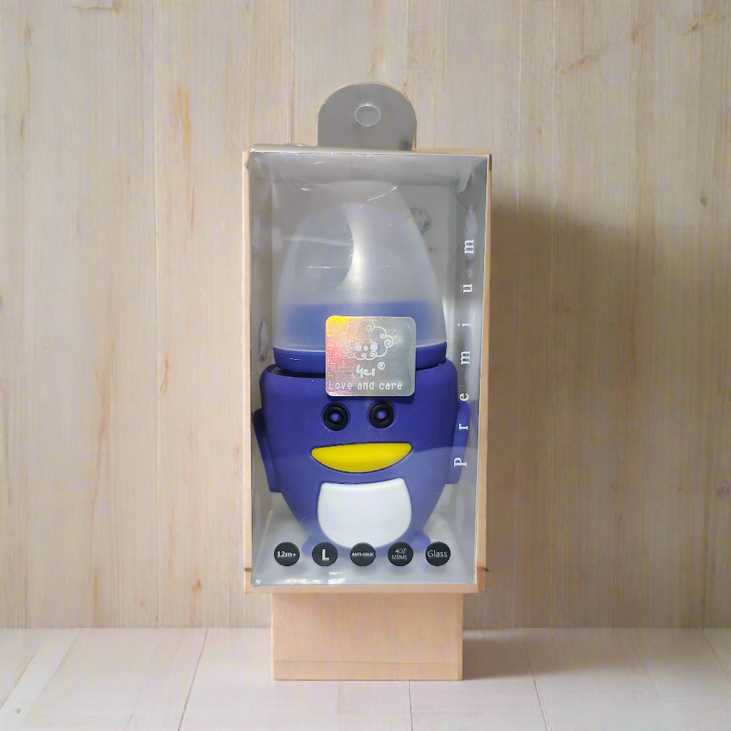 Blue penguin-shaped baby bottle in clear packaging on a white surface with a blurred background.