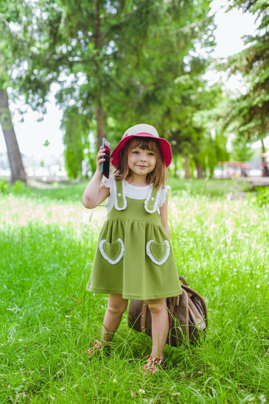 Young girl in a green dress with heart pockets and a red hat standing in a grassy field.