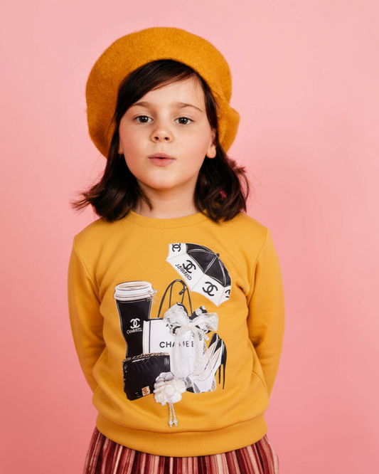 Child wearing a mustard yellow sweatshirt with graphic design on a pink background