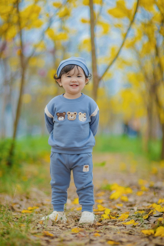 Child in a blue outfit with bear designs standing in a park with yellow leaves.