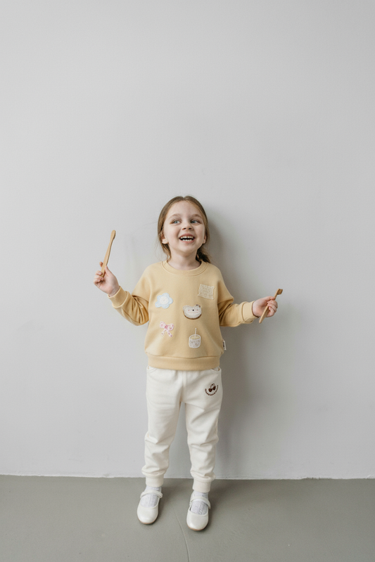 Child wearing a yellow sweater with decorative elements and white pants, standing against a plain wall.